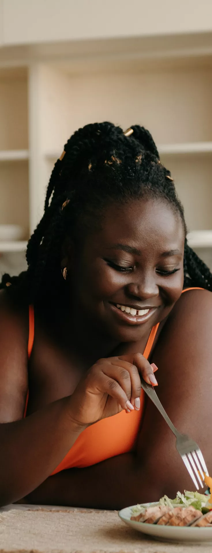 A woman sits at a kitchen table, eating a fresh salad with a fork. The setting is a bright, modern kitchen with open shelving and neutral tones. A glass of orange juice and a bottle are visible on the table, adding to the casual dining atmosphere. 
