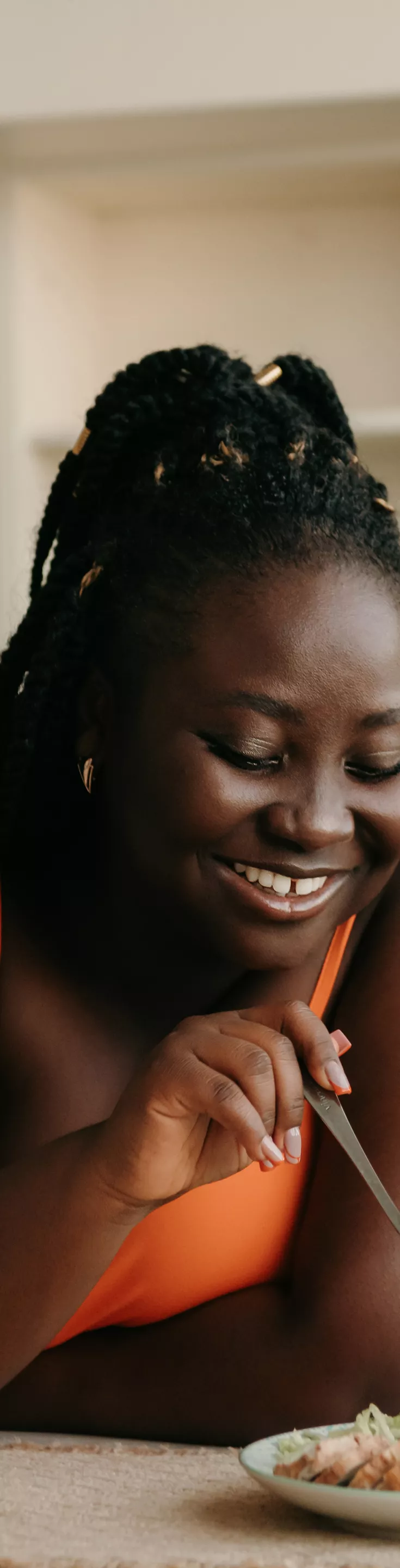 A woman sits at a kitchen table, eating a fresh salad with a fork. The setting is a bright, modern kitchen with open shelving and neutral tones. A glass of orange juice and a bottle are visible on the table, adding to the casual dining atmosphere. 