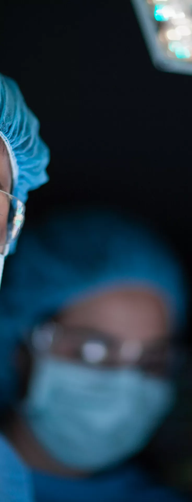 A medical professional dressed in blue surgical scrubs and a cap stands under bright operating room lights. The setting is a modern surgical theater, with focus on the illuminated environment and sterile attire. 