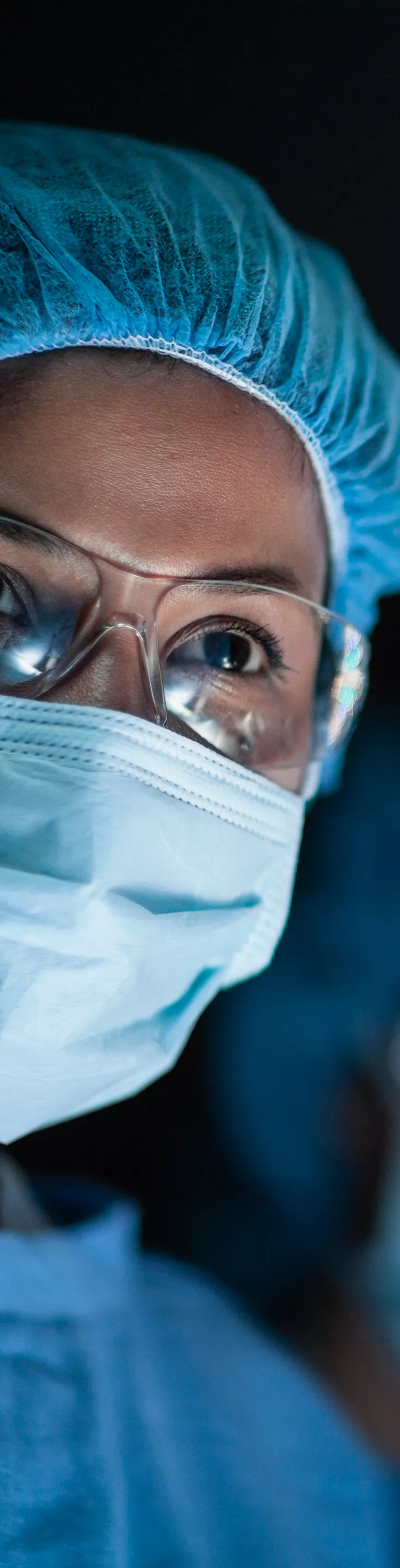 A medical professional dressed in blue surgical scrubs and a cap stands under bright operating room lights. The setting is a modern surgical theater, with focus on the illuminated environment and sterile attire. 