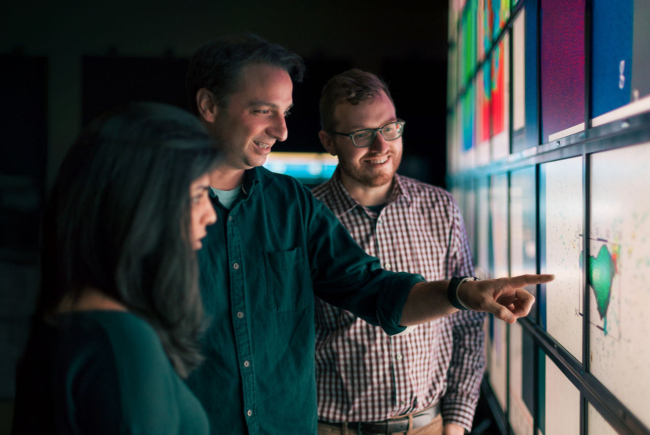 A group of three adults stands in front of a large illuminated digital display wall, analyzing and discussing the content. The setting appears to be a modern office or creative workspace, with one person actively pointing at the screen.