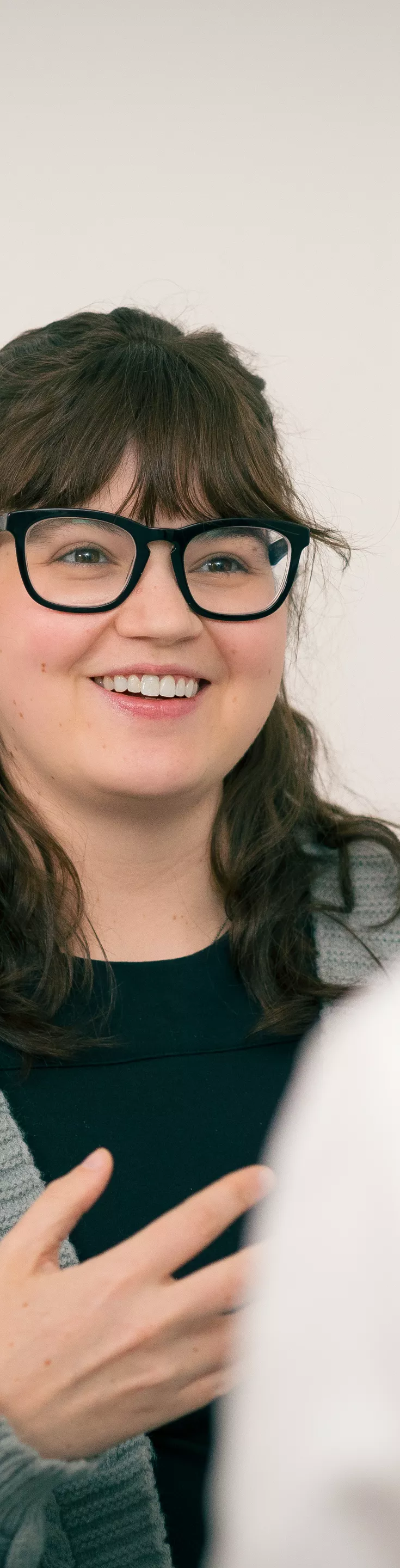 The image shows two adults facing each other in a neutral indoor setting. One person, wearing a gray sweater, gestures with their hand while speaking, suggesting a serious or important discussion. The other individual, seen from behind, wears a white garment, possibly a coat. 