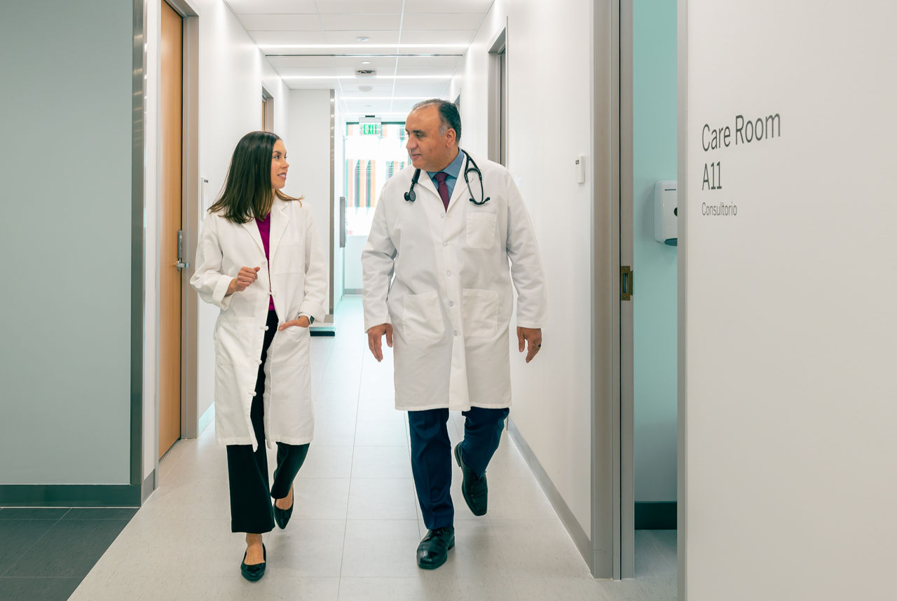 A male and female doctor walk together down a brightly lit hospital hallway. Both are dressed in white lab coats, with the male doctor wearing a stethoscope. The setting is clean and modern, with a visible sign reading 'Care Room A11' on the wall.
