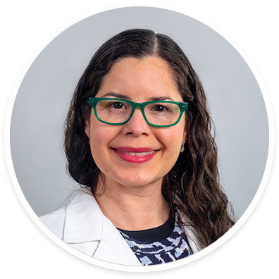 A woman with long, wavy dark hair is pictured in a studio setting. She is wearing a white coat over a patterned top, suggesting a professional or medical context. The background is a plain, light gray, creating a neutral and clean visual. The image is presented in a circular frame.