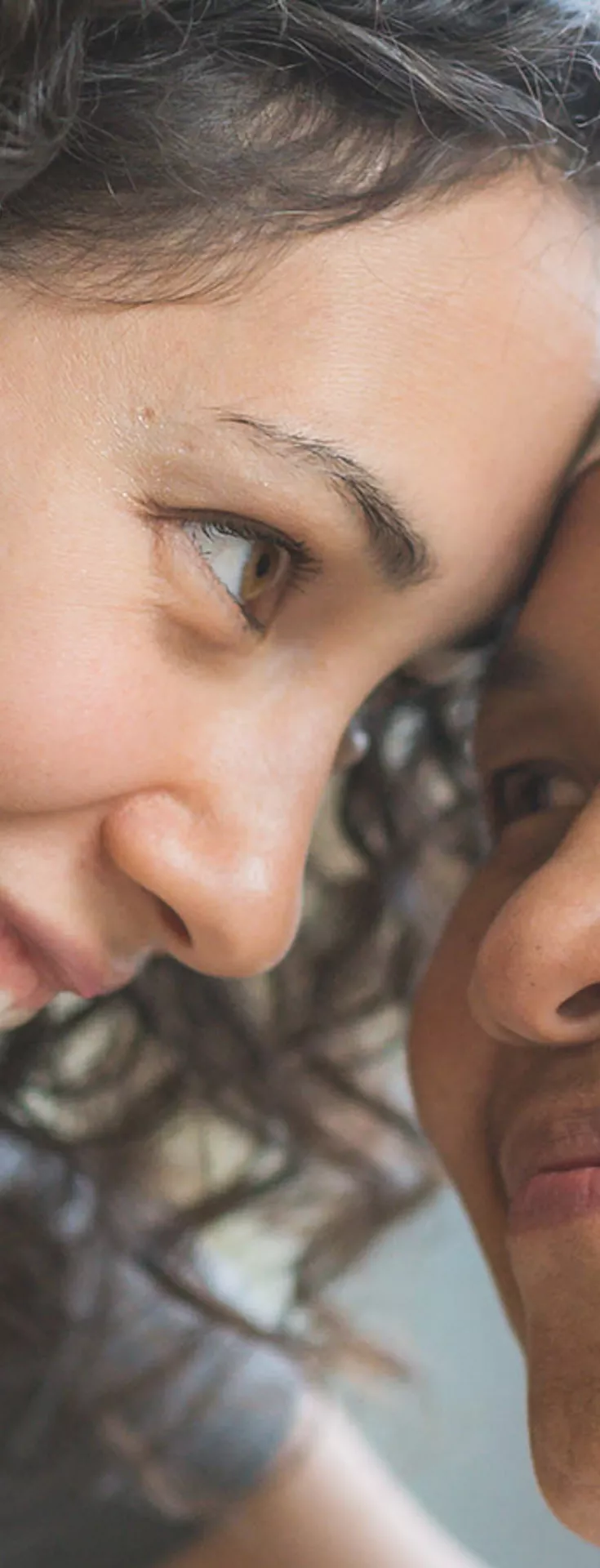 Two women are seated closely together in an indoor setting, one wearing a patterned headscarf. The atmosphere appears caring and intimate, with soft natural light coming through a window in the background.