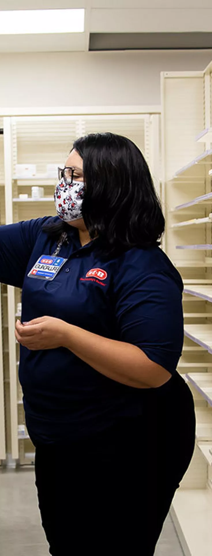 A woman wearing a mask and navy shirt is seen arranging bottles in a pharmacy aisle. The setting is a well-lit, organized pharmacy with rows of white medicine containers. 