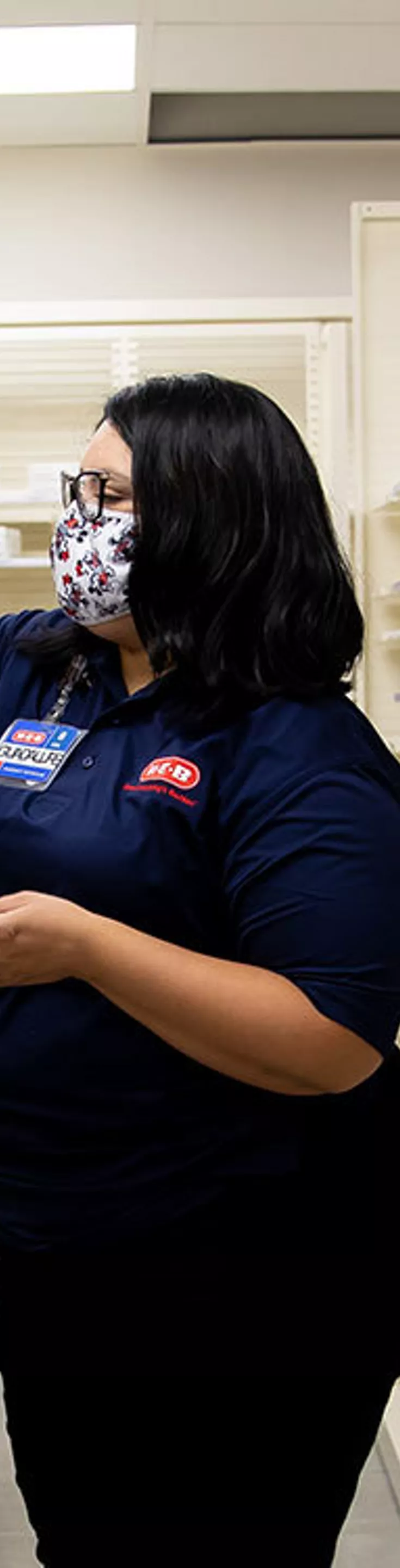A woman wearing a mask and navy shirt is seen arranging bottles in a pharmacy aisle. The setting is a well-lit, organized pharmacy with rows of white medicine containers. 