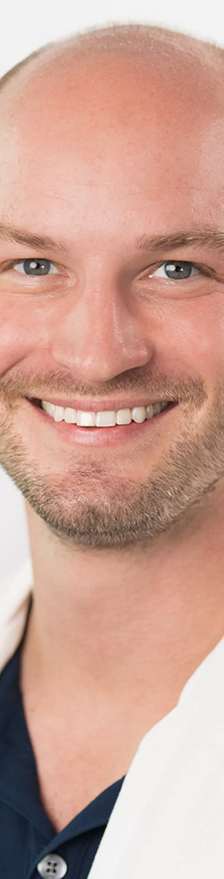 Headshot of Branden Daubel. He is wearing a navy dress shirt and a white coat.
