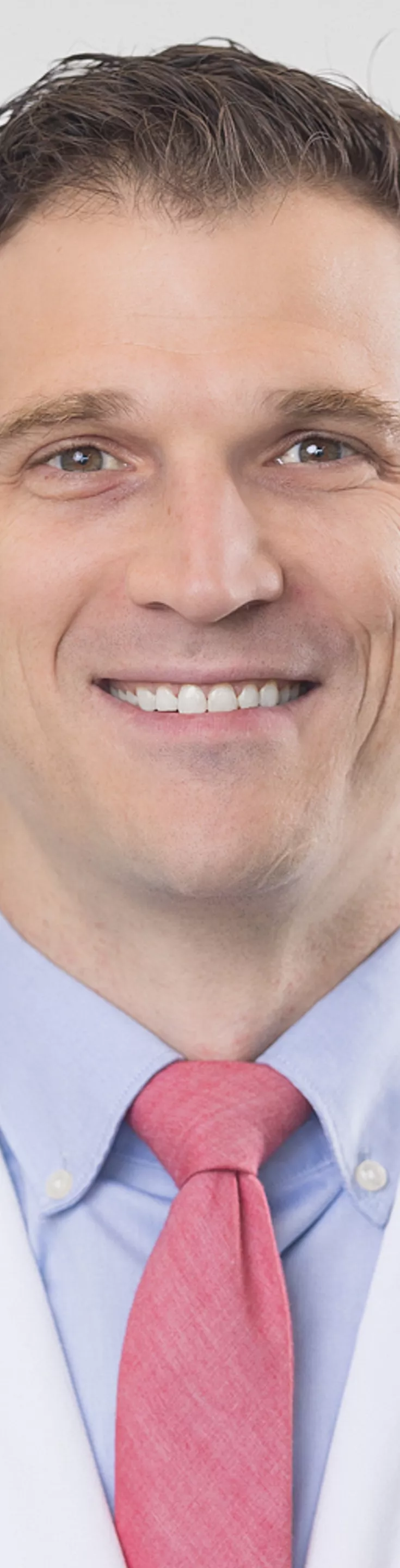 Headshot of John Wickman. He is wearing a blue dress shirt, red tie, and a white coat.