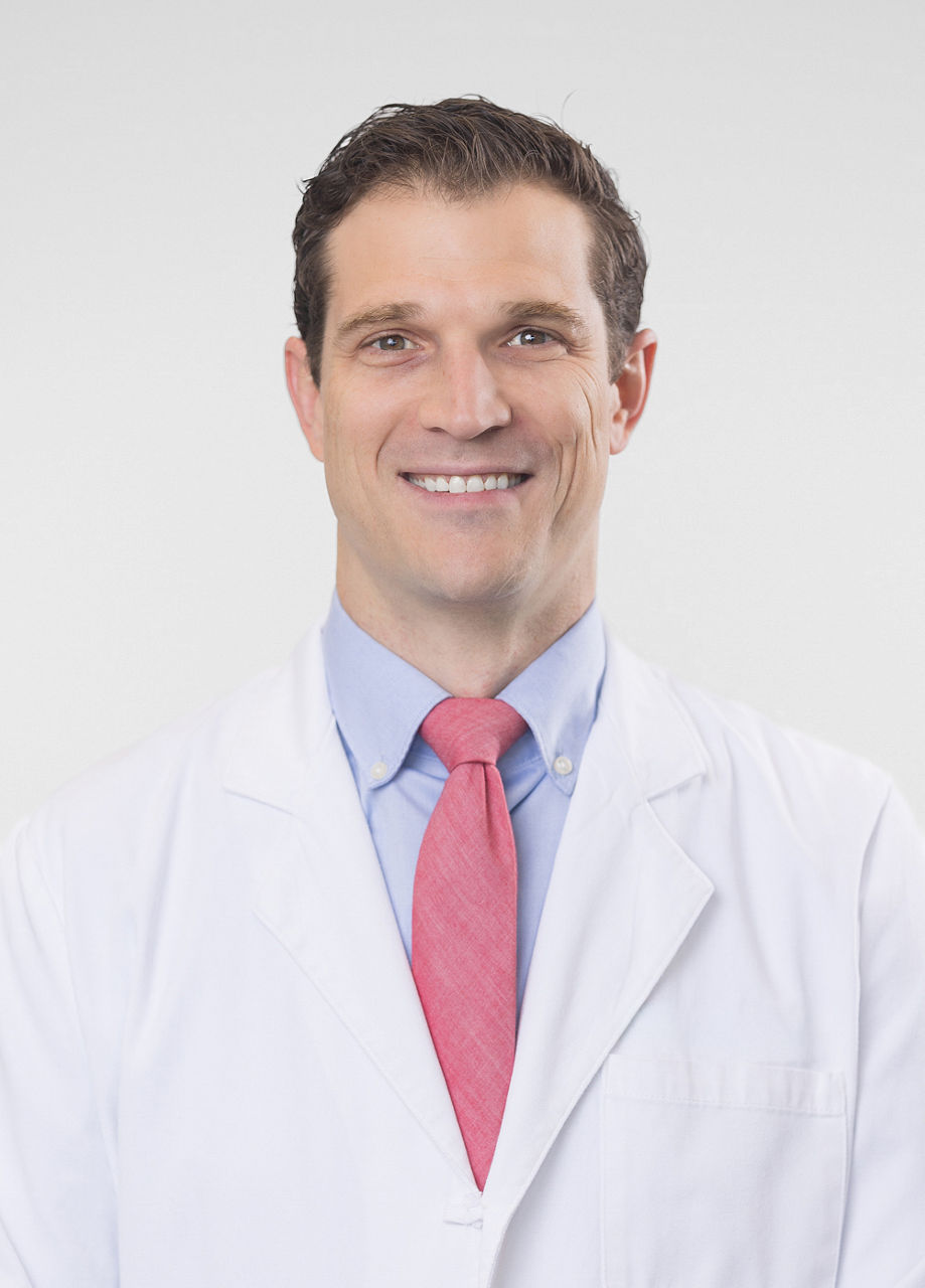 Headshot of John Wickman. He is wearing a blue dress shirt, red tie, and a white coat.