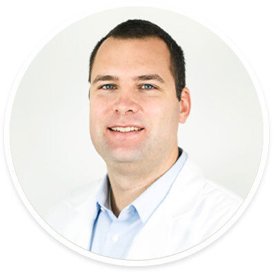 A man is pictured wearing a white lab coat over a light blue collared shirt. The background is plain and white, emphasizing a clean and clinical setting. The image is cropped in a circular frame, focusing on the upper body and attire. The overall mood is professional and neutral.