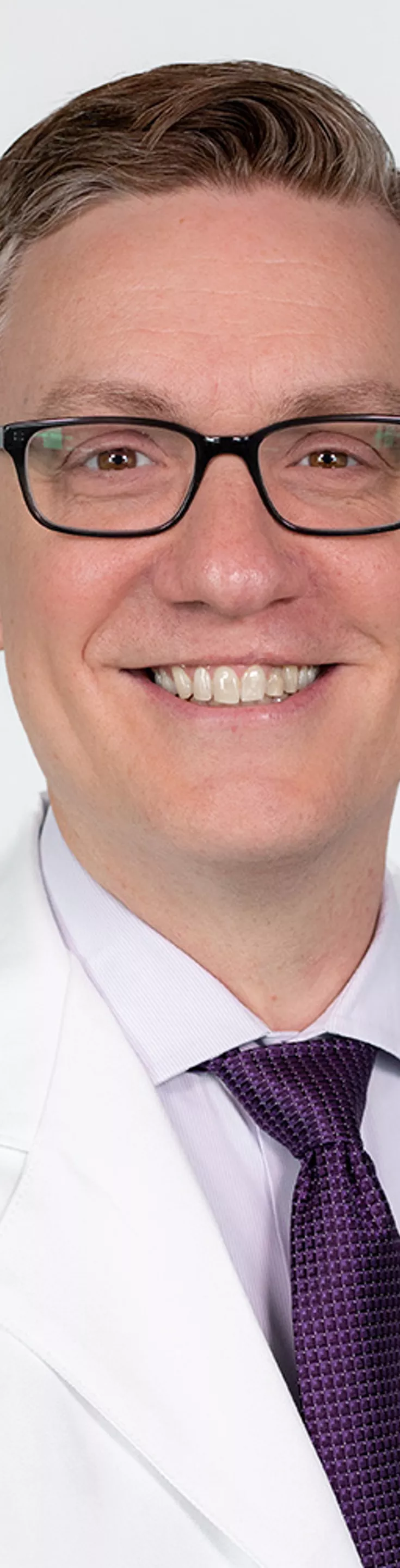 Headshot of Steve Steffensen. He is wearing a dress shirt, purple tie, and a white coat.