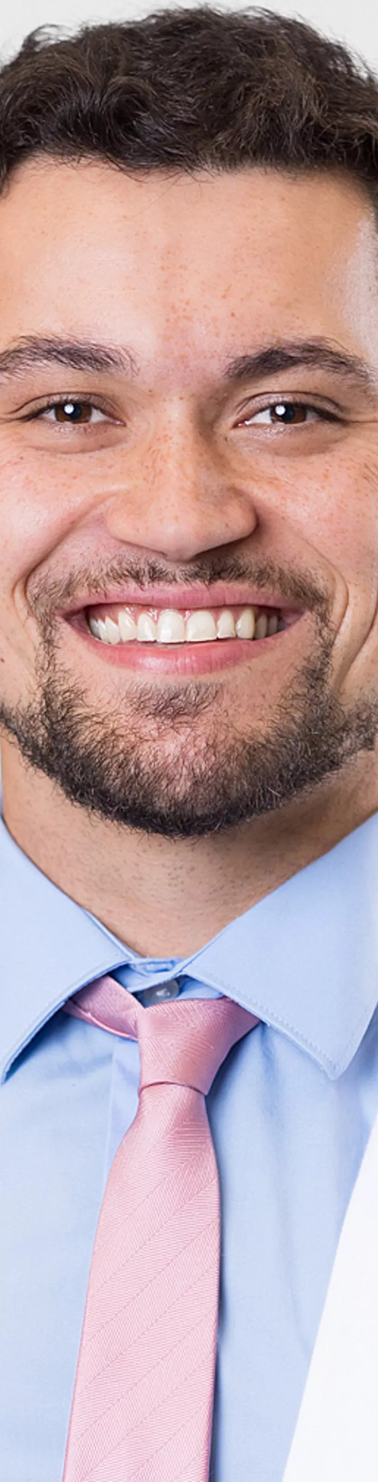 Headshot of Thomas Ratana. He is wearing a blue dress shirt, a pink tie, and a white coat.