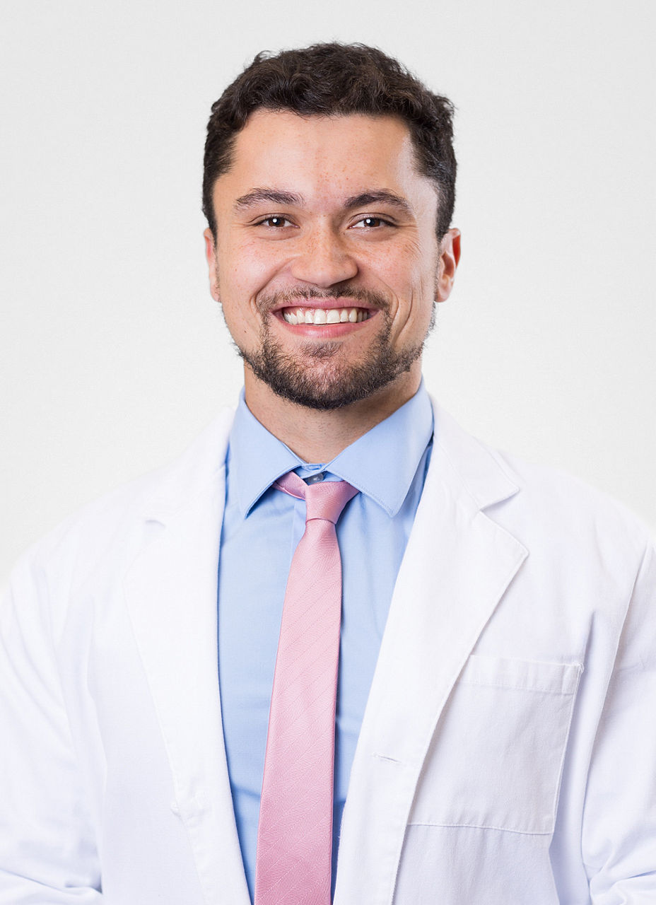 Headshot of Thomas Ratana. He is wearing a blue dress shirt, a pink tie, and a white coat.