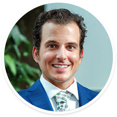 A man is dressed in a sharp blue suit paired with a white shirt and a floral-patterned tie. The background features green foliage, suggesting an outdoor or garden setting. The image is cropped in a circular frame, focusing on the upper body and attire. The overall mood is formal and professional, with a touch of style from the patterned tie.