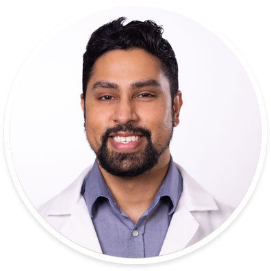 A person is shown wearing a white lab coat over a blue collared shirt. The background is plain white, creating a clean and clinical setting. The subject's attire suggests a professional or medical context. The image is well-lit with neutral tones, emphasizing a professional and formal mood.