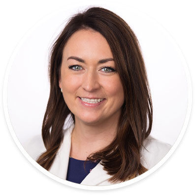 A woman with medium-length brown hair is pictured in a studio setting. She is wearing a white blazer over a dark top, standing against a plain white background. The image is framed in a circular crop, giving it a clean and modern look. The overall style is professional and suitable for business or corporate use.
