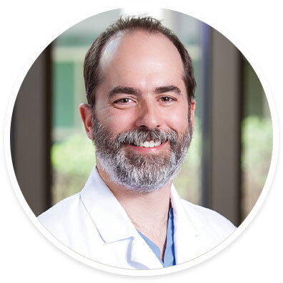 A person is shown wearing a white lab coat, suggesting a medical or healthcare professional. The background features blurred greenery and large windows, indicating an outdoor or hospital setting. The subject's attire includes a blue shirt or scrubs under the coat. The image is presented in a circular frame.