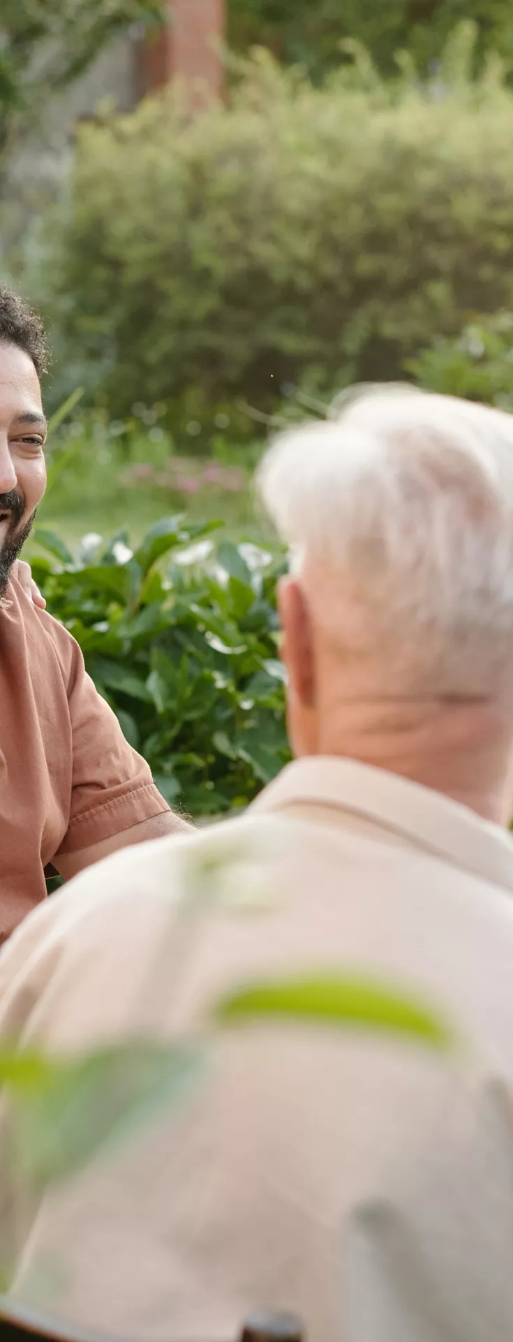 A group of people are seated around a table outdoors, sharing a meal in a lush garden setting. The scene includes adults and a child, suggesting a multigenerational family gathering. The table is set with plates of food, drinks, and fresh greenery, creating a warm and inviting atmosphere. 