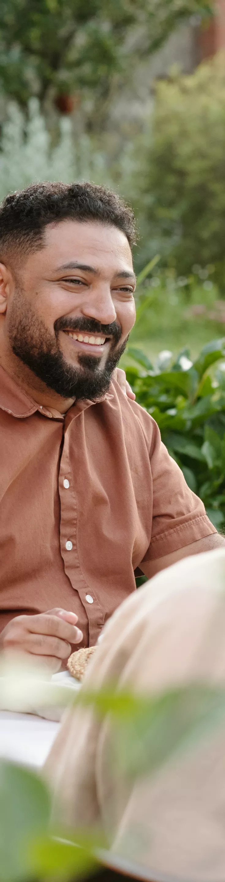 A group of people are seated around a table outdoors, sharing a meal in a lush garden setting. The scene includes adults and a child, suggesting a multigenerational family gathering. The table is set with plates of food, drinks, and fresh greenery, creating a warm and inviting atmosphere. 