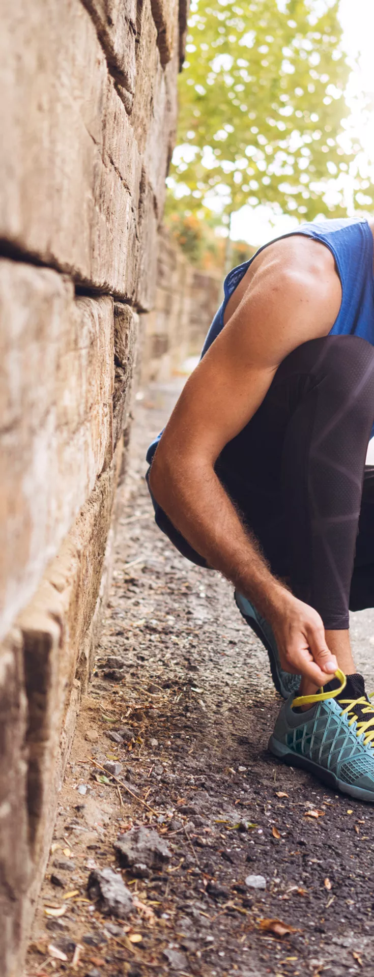 A man in athletic wear is crouched down on a paved path, tying his blue running shoes. Beside him sits a cheerful yellow Labrador Retriever, looking up attentively. The setting is an outdoor park or trail with trees and a stone wall, suggesting a casual exercise or jogging activity. 