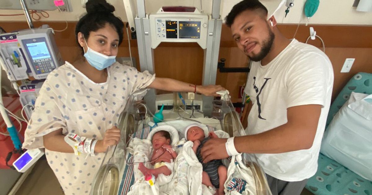 Two newborn babies are lying in a hospital neonatal incubator, each wrapped in blankets. Medical equipment and monitors are visible in the background, indicating a clinical setting. Two adults stand on either side of the incubator, suggesting a family moment in the hospital. The environment is bright and sterile, with a focus on the infants' care. Hospital wristbands and patient monitors add to the medical context.