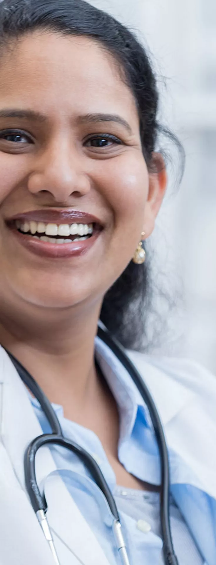 A woman wearing a white lab coat and stethoscope is present in a clinical laboratory environment. The background shows another person in medical attire and laboratory equipment, suggesting an active healthcare or research setting. The image features bright, clinical lighting and a professional atmosphere. 