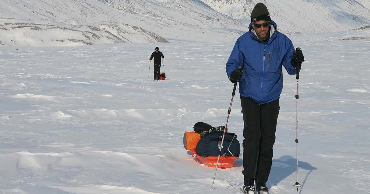 Two individuals are trekking across a vast, snow-covered terrain, each pulling a loaded sled behind them. The person in the foreground wears a blue jacket, dark pants, and a hat, using ski poles for support. The background features another trekker and expansive snowfields with distant hills. The scene conveys a sense of adventure and endurance in a cold, remote environment.