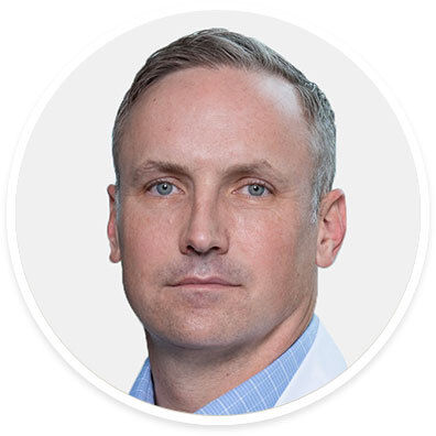 A man with short, grayish hair is pictured wearing a light blue checkered shirt and a white coat. The background is plain and white, creating a clean and professional look. The image is cropped in a circular frame, focusing on the upper torso and head. The overall style is formal and suitable for business or medical contexts.