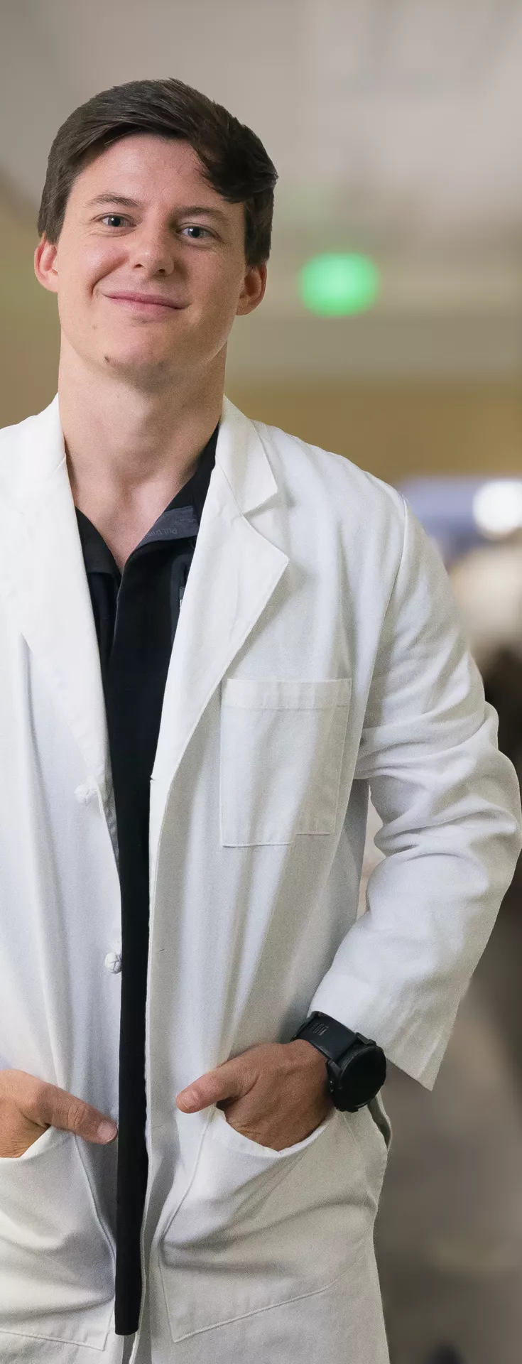 A male optometrist stands confidently in a modern eye clinic, wearing a white lab coat. He is positioned next to specialized ophthalmic equipment, likely used for eye examinations. The clinic setting is bright and professional, with a clean and organized workspace visible in the background. 