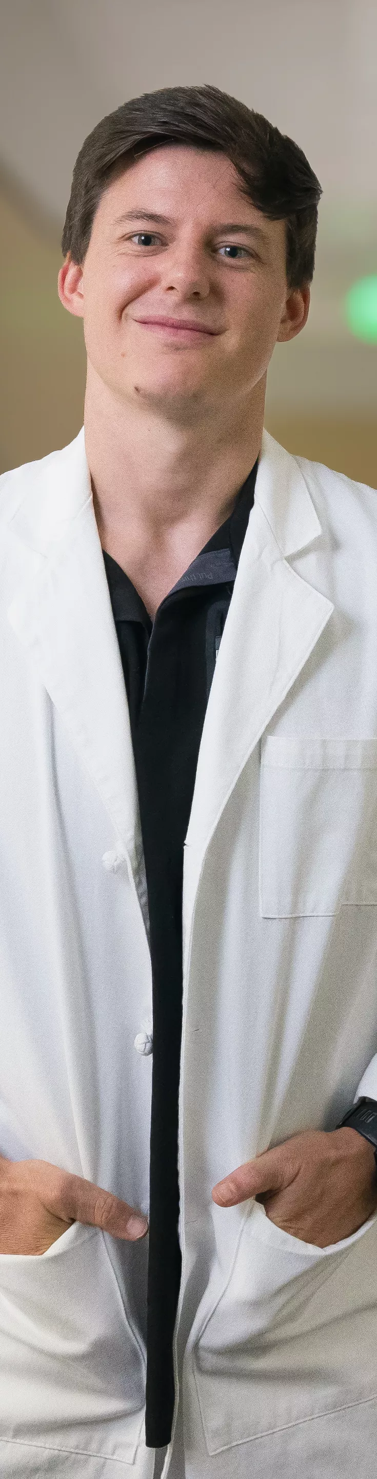 A male optometrist stands confidently in a modern eye clinic, wearing a white lab coat. He is positioned next to specialized ophthalmic equipment, likely used for eye examinations. The clinic setting is bright and professional, with a clean and organized workspace visible in the background. 