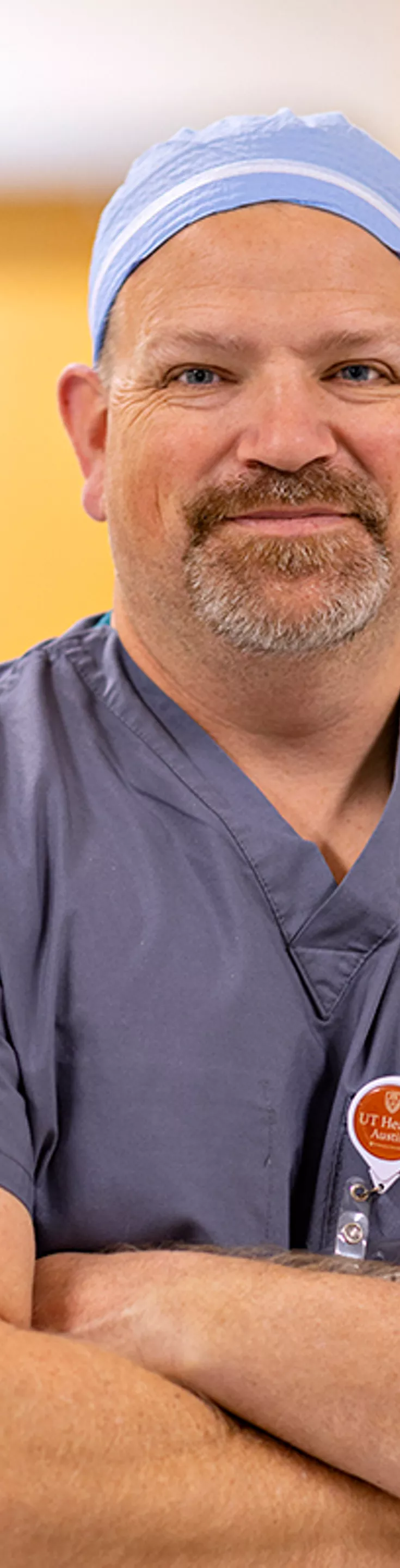 A person wearing blue-grey medical scrubs and a cap stands with arms crossed in a brightly lit hospital hallway. The individual has a visible ID badge clipped to their chest and holds a white face mask. The background features out-of-focus double doors, suggesting a clinical or healthcare environment.