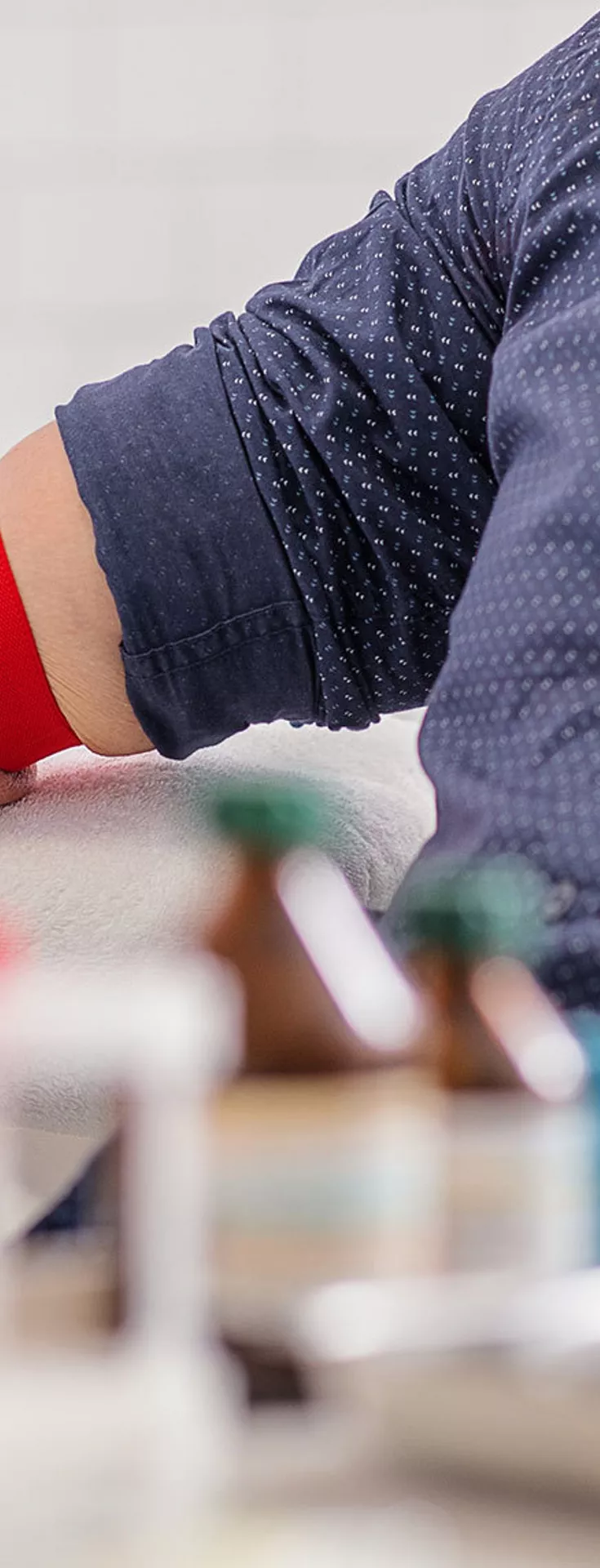 A healthcare professional wearing blue gloves is preparing an adult male patient for a blood draw by applying a red tourniquet to his arm. The setting appears to be a clinical laboratory or medical office, with test tubes and medical supplies visible in the foreground. 