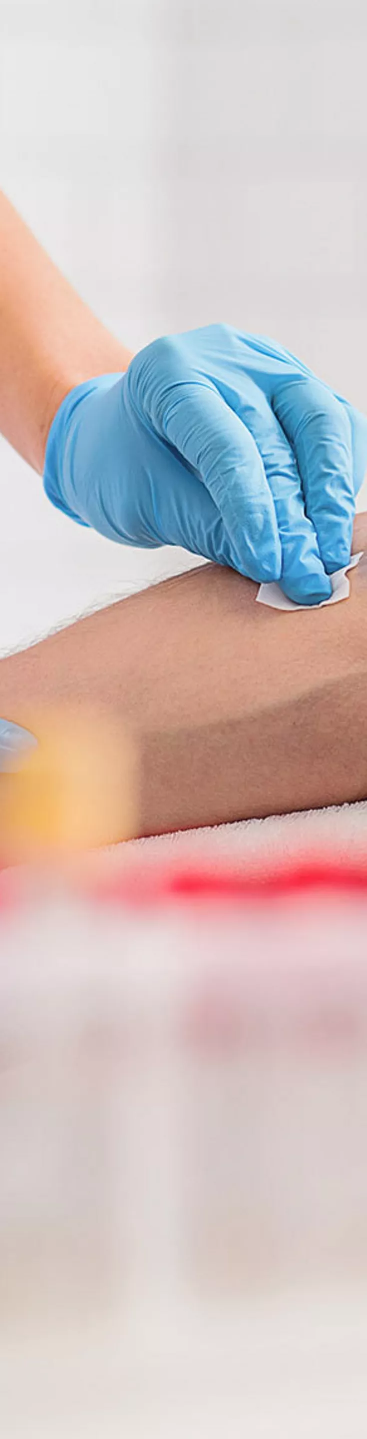 A healthcare professional wearing blue gloves is preparing an adult male patient for a blood draw by applying a red tourniquet to his arm. The setting appears to be a clinical laboratory or medical office, with test tubes and medical supplies visible in the foreground. 