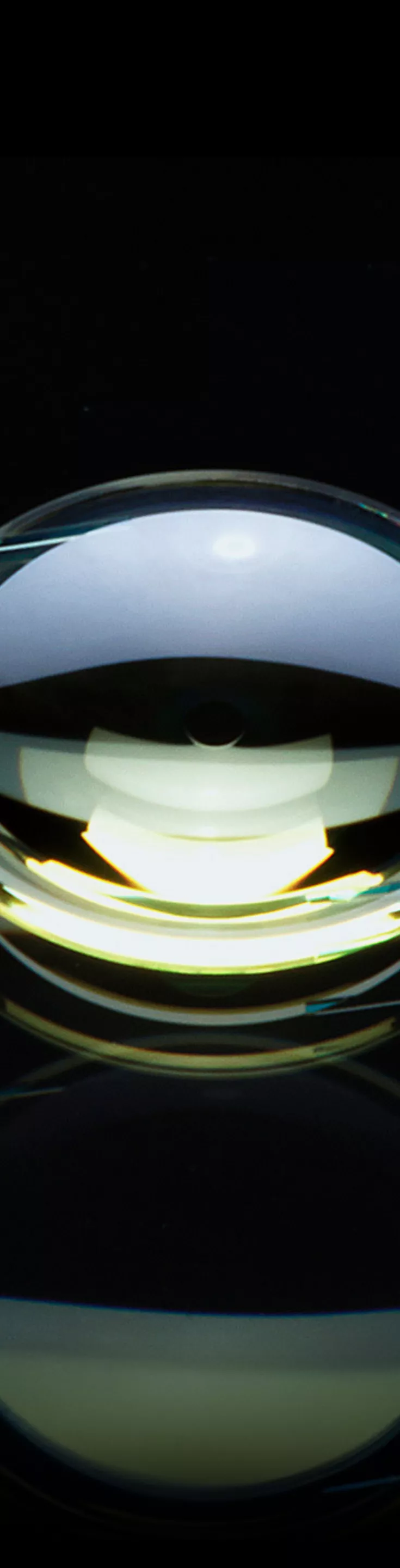 A single intraocular lens is displayed against a glossy black surface, highlighting its transparent, curved structure and fine supporting arms. The lens reflects ambient light, creating a dramatic and clinical visual effect. 