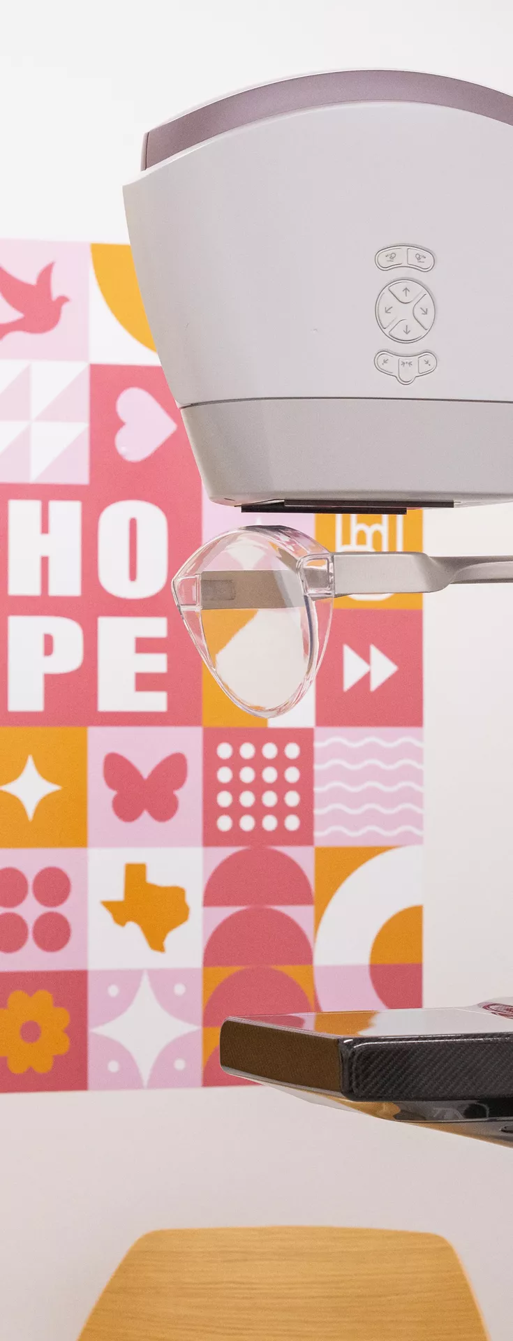 A contemporary mammography device is positioned in a clinical setting, with a colorful wall art featuring the word 'HOPE' and various pink-themed icons in the background. The machine displays clear medical technology details, and the environment suggests a healthcare facility focused on breast cancer awareness. 
