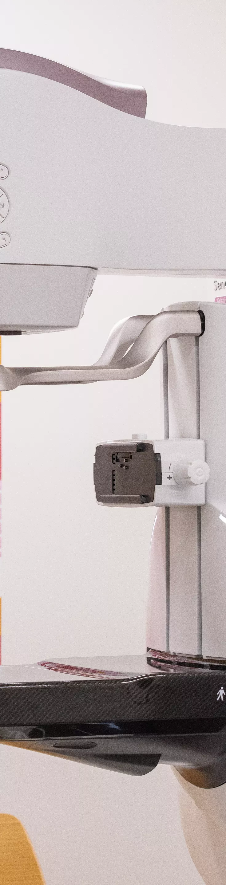 A contemporary mammography device is positioned in a clinical setting, with a colorful wall art featuring the word 'HOPE' and various pink-themed icons in the background. The machine displays clear medical technology details, and the environment suggests a healthcare facility focused on breast cancer awareness. 