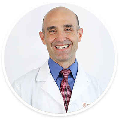 A male adult is shown wearing a white medical coat over a blue dress shirt and a red patterned tie. The setting is a plain, light background, emphasizing a professional and clinical environment. The attire and context suggest a healthcare or medical professional. No visible text, numbers, or identifiable logos are present in the image.