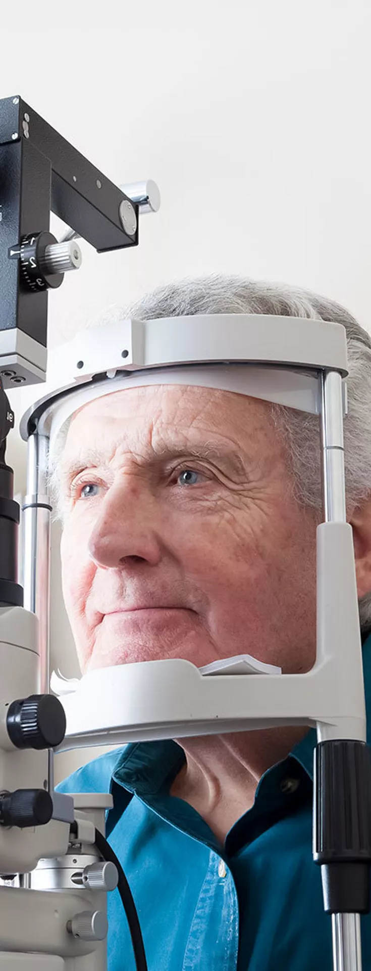 An older man is seated at an ophthalmology clinic, having his eyes examined with a slit lamp device. A healthcare professional is conducting the eye test in a clinical setting with neutral walls. 