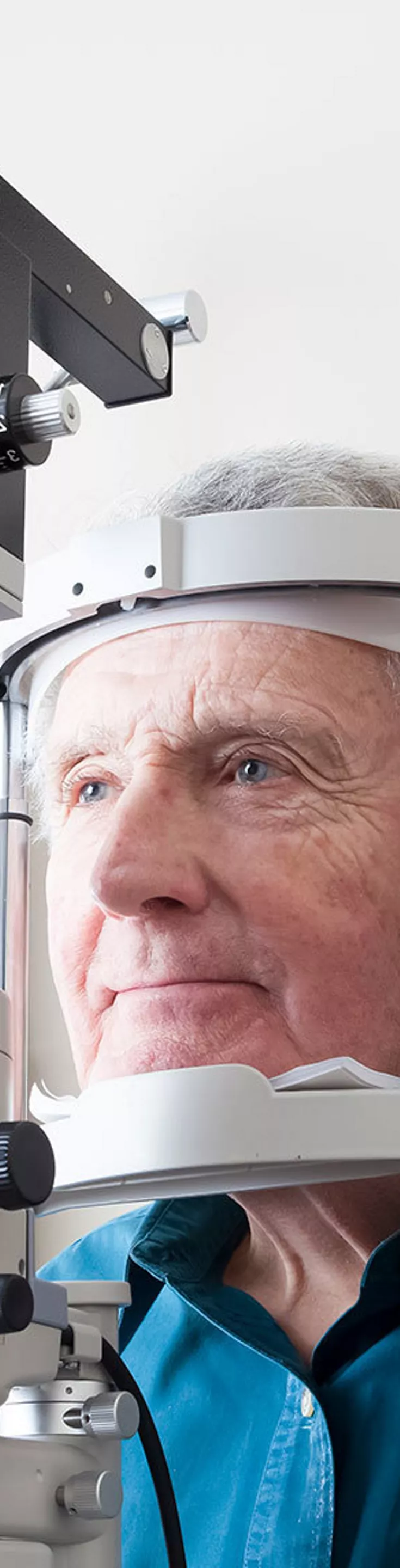 An older man is seated at an ophthalmology clinic, having his eyes examined with a slit lamp device. A healthcare professional is conducting the eye test in a clinical setting with neutral walls. 