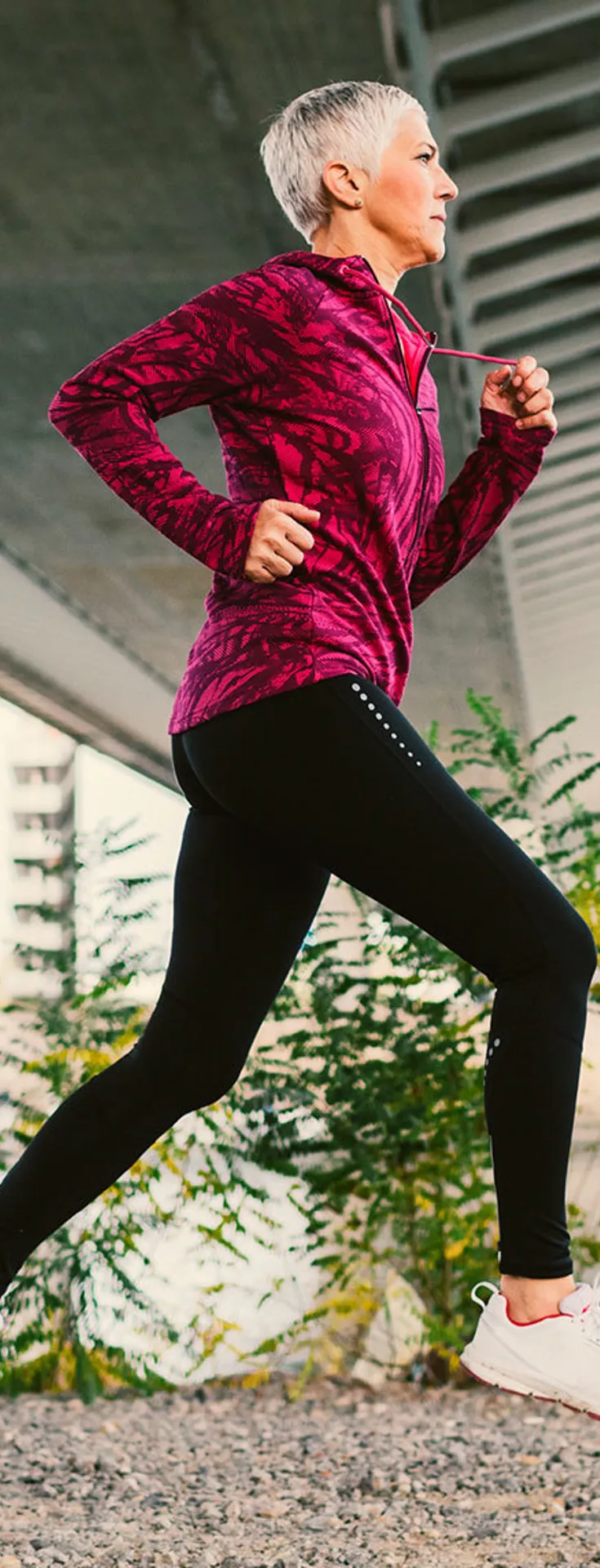 A senior woman with short gray hair is jogging on a gravel path beneath a large urban bridge. She wears a bright patterned jacket and black leggings, moving energetically in an outdoor setting with greenery and water in the background. 