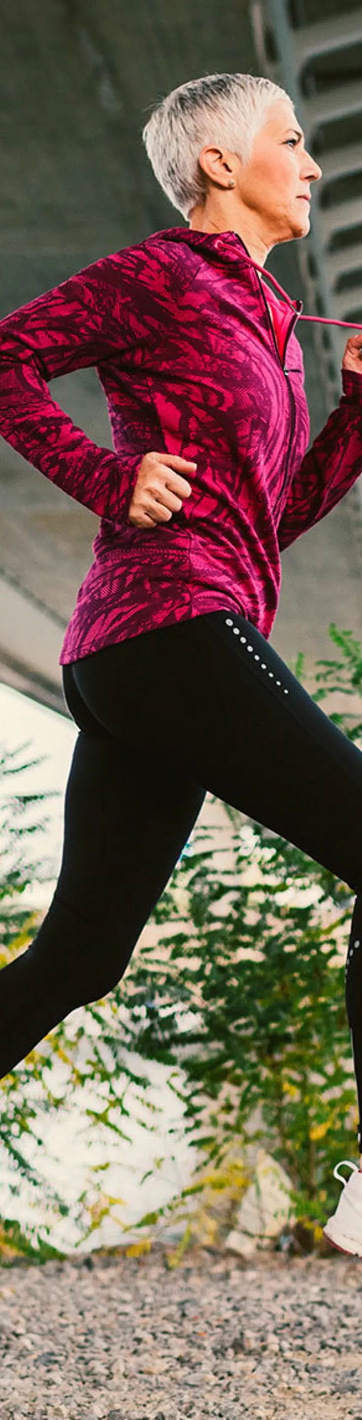 A senior woman with short gray hair is jogging on a gravel path beneath a large urban bridge. She wears a bright patterned jacket and black leggings, moving energetically in an outdoor setting with greenery and water in the background. 