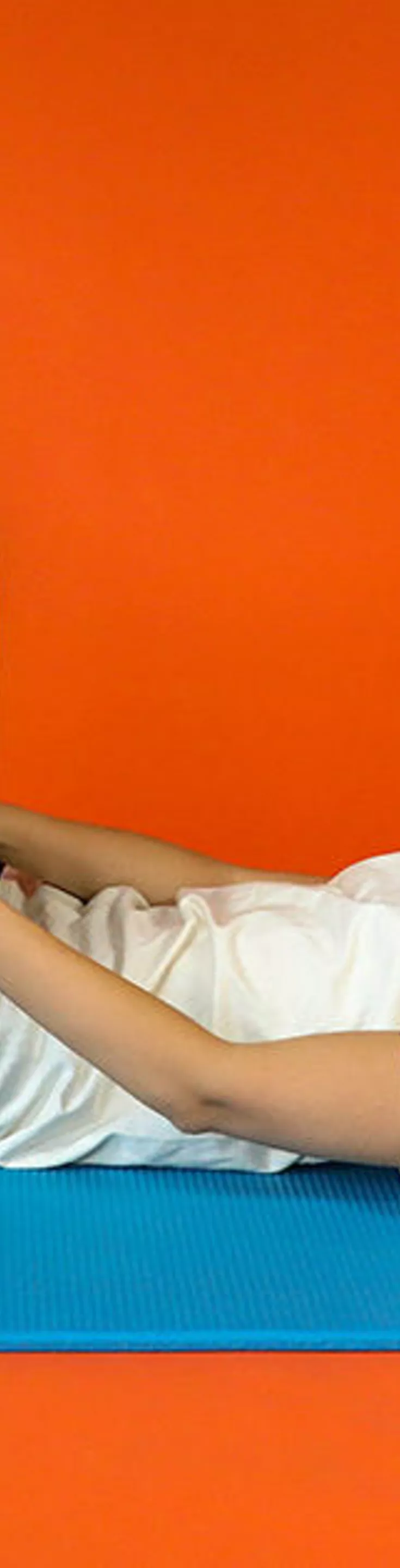 A young woman lies on her back on a blue exercise mat, performing a knee-to-chest stretch. She is dressed in a white t-shirt and black leggings, set against a solid orange backdrop. The scene conveys a calm and focused mood, emphasizing fitness and flexibility. No visible text or numbers are present in the image.