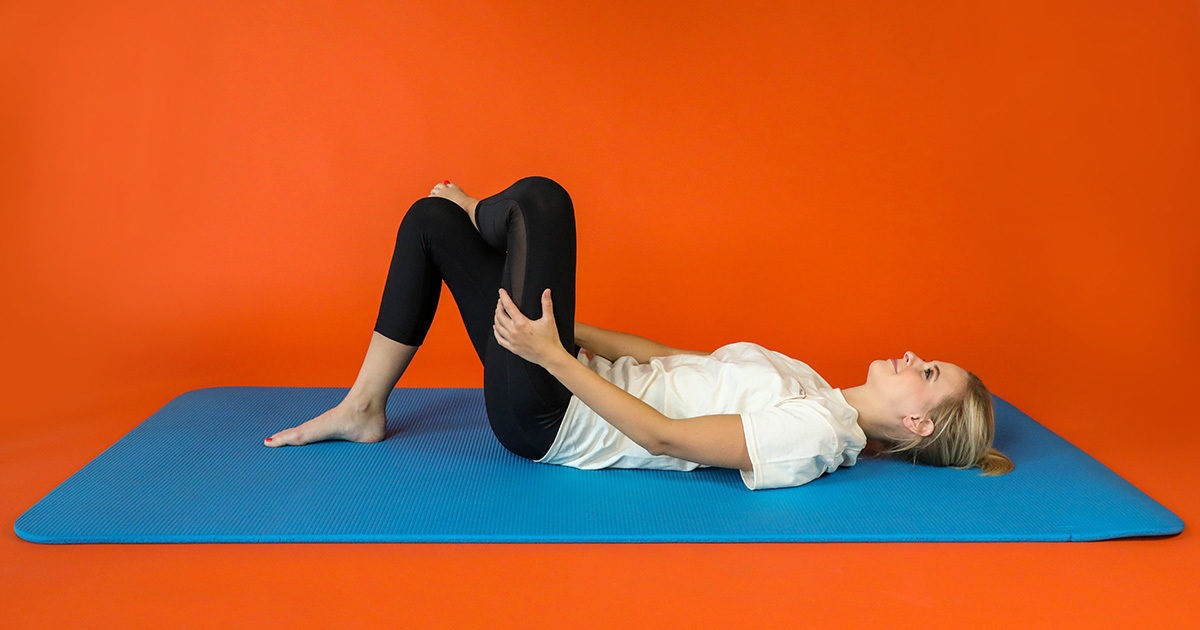 A young woman lies on her back on a blue exercise mat, performing a knee-to-chest stretch. She is dressed in a white t-shirt and black leggings, set against a solid orange backdrop. The scene conveys a calm and focused mood, emphasizing fitness and flexibility. No visible text or numbers are present in the image.
