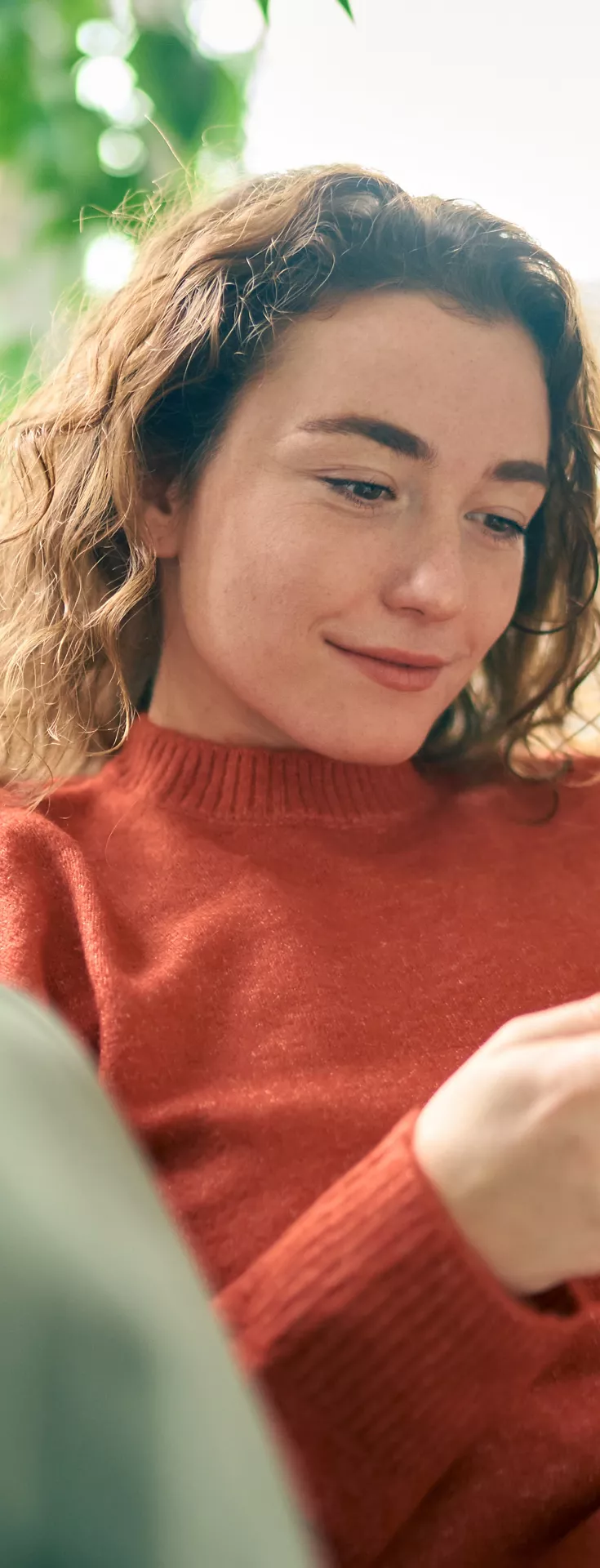 A young woman sits comfortably on a sofa, focused on her yellow smartphone. The setting is a cozy, well-lit living room with natural light streaming through the window. She wears a casual red sweater, and the atmosphere feels relaxed and informal. 