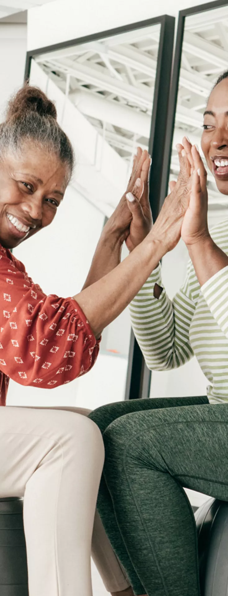 Two women are seated on large stability balls, facing each other and engaging in a playful or fitness activity. The setting is a bright, modern studio with mirrors and natural light.