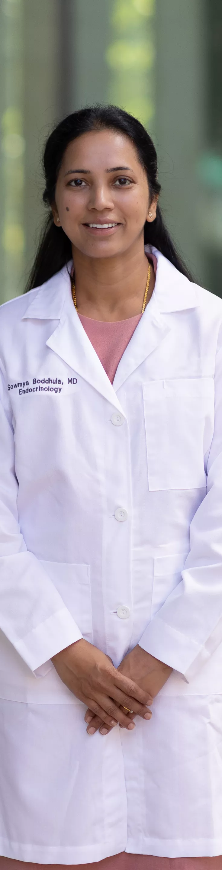 A woman stands outside wearing a white lab coat over a pink dress. The setting appears to be an urban or institutional environment with glass and concrete elements visible in the background. 