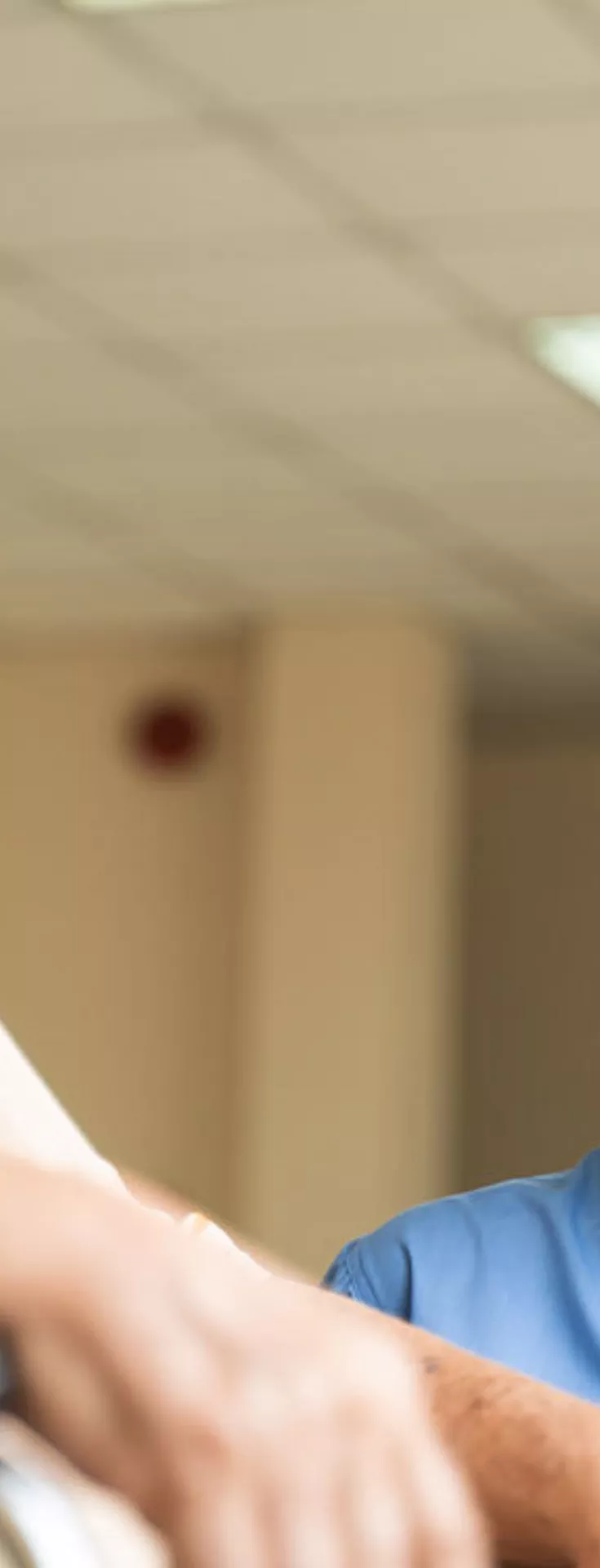 A healthcare worker in blue scrubs is attending to an elderly person seated in a wheelchair. The setting appears to be a well-lit indoor facility, possibly a hospital or care home, with a tiled ceiling and fluorescent lighting. The nurse is wearing a face mask, indicating a focus on health and safety. The interaction suggests care, support, and attentive service.