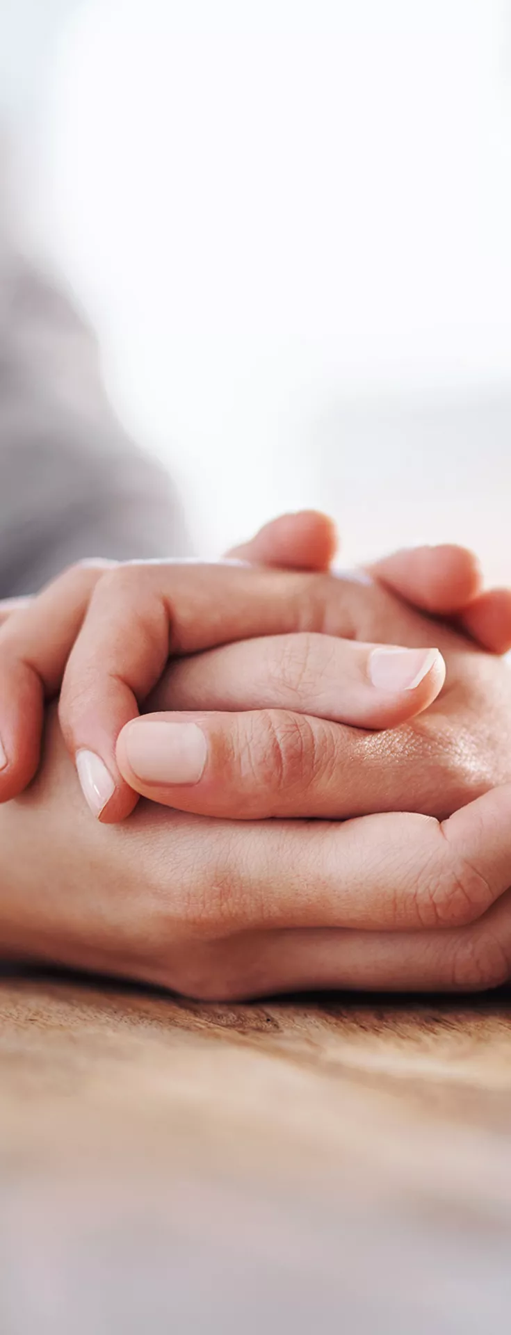 The main subject is a close-up of two people holding hands, conveying support and empathy. The setting appears to be indoors, with a wooden table surface visible. 