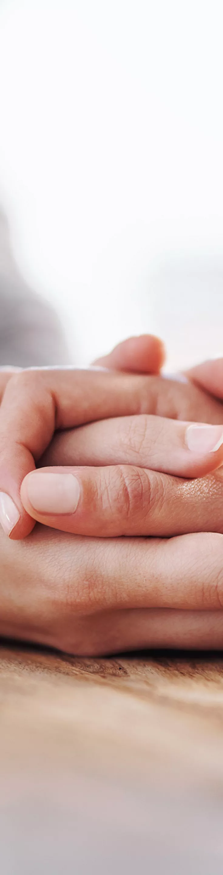 The main subject is a close-up of two people holding hands, conveying support and empathy. The setting appears to be indoors, with a wooden table surface visible. 